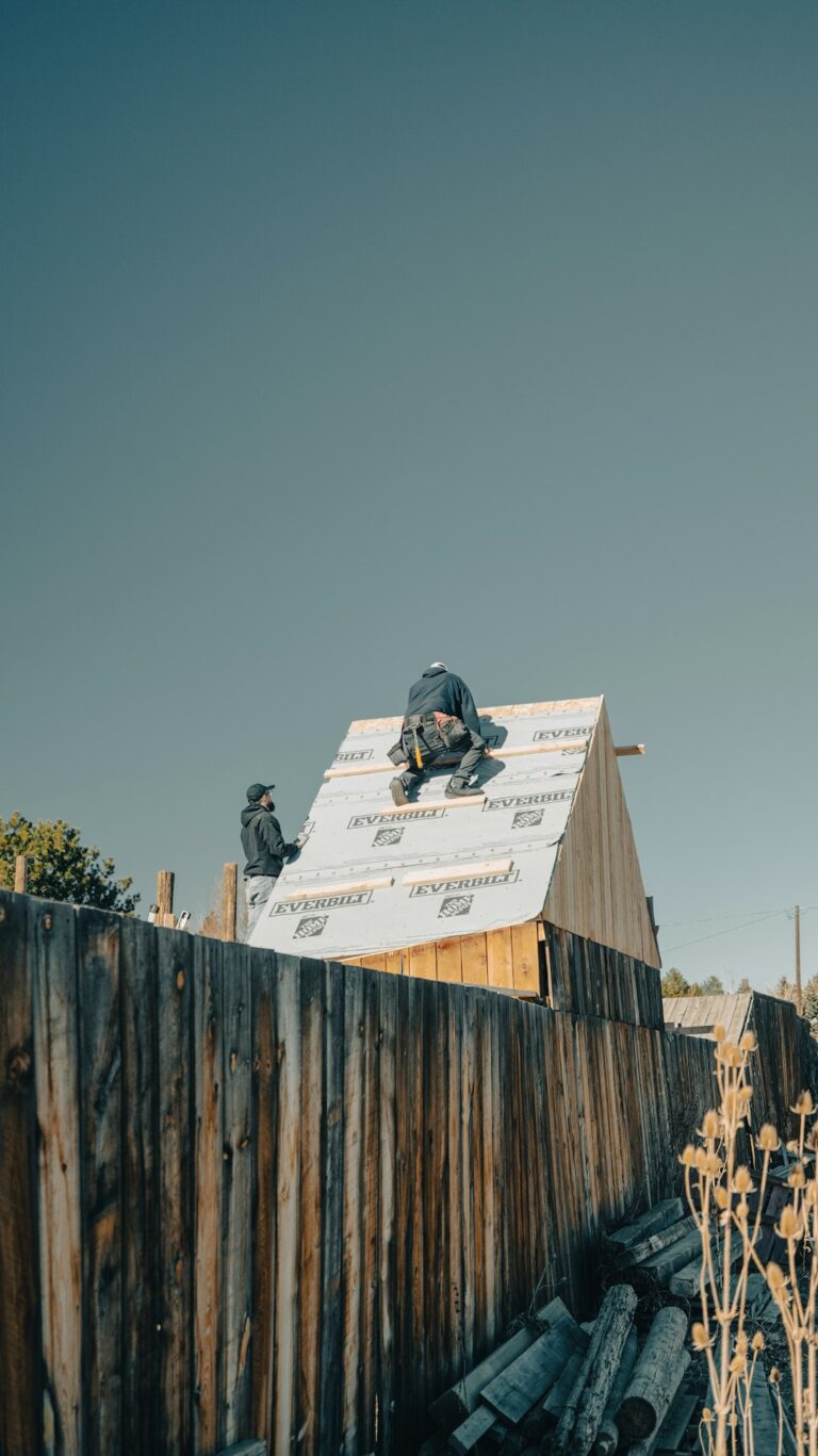 Workers secure a weatherproof tarp over an exposed roof section beside renovation materials and a ladder.